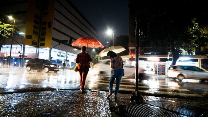 Pessoas com guarda-chuva caminham sob chuva em rua de cidade brasileira